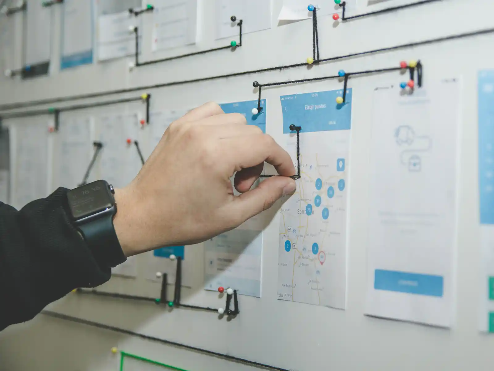 A hand connecting printed app screens pinned to a wall with string, mapping out a user journey
