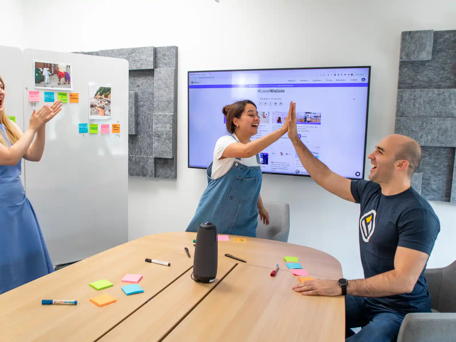 Three colleagues celebrating with a high five in a meeting room, a website shown on the screen behind them